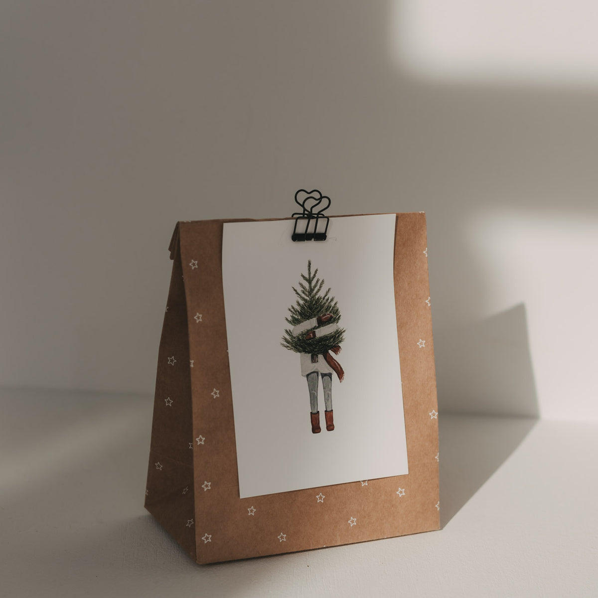 Schwarzer Herz-Clip hält eine Weihnachtskarte mit Tannenbaum-Motiv an einer braunen Geschenktüte mit Sternen.