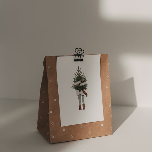Schwarzer Herz-Clip hält eine Weihnachtskarte mit Tannenbaum-Motiv an einer braunen Geschenktüte mit Sternen.
