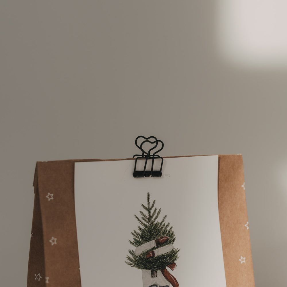 Nahaufnahme eines schwarzen Foldback-Clips mit Herzmotiv, der eine Weihnachtskarte an einer Geschenktüte befestigt.