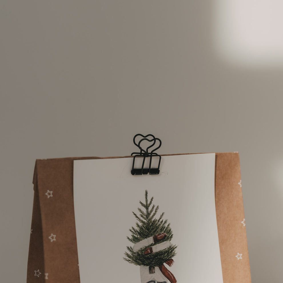 Nahaufnahme eines schwarzen Foldback-Clips mit Herzmotiv, der eine Weihnachtskarte an einer Geschenktüte befestigt.
