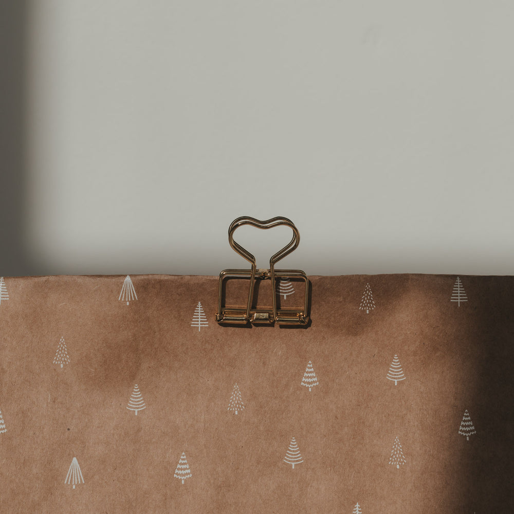 Goldener Herz-Clip von Eulenschnitt verschließt eine braune Geschenktüte mit weißem Tannenbaum-Muster.