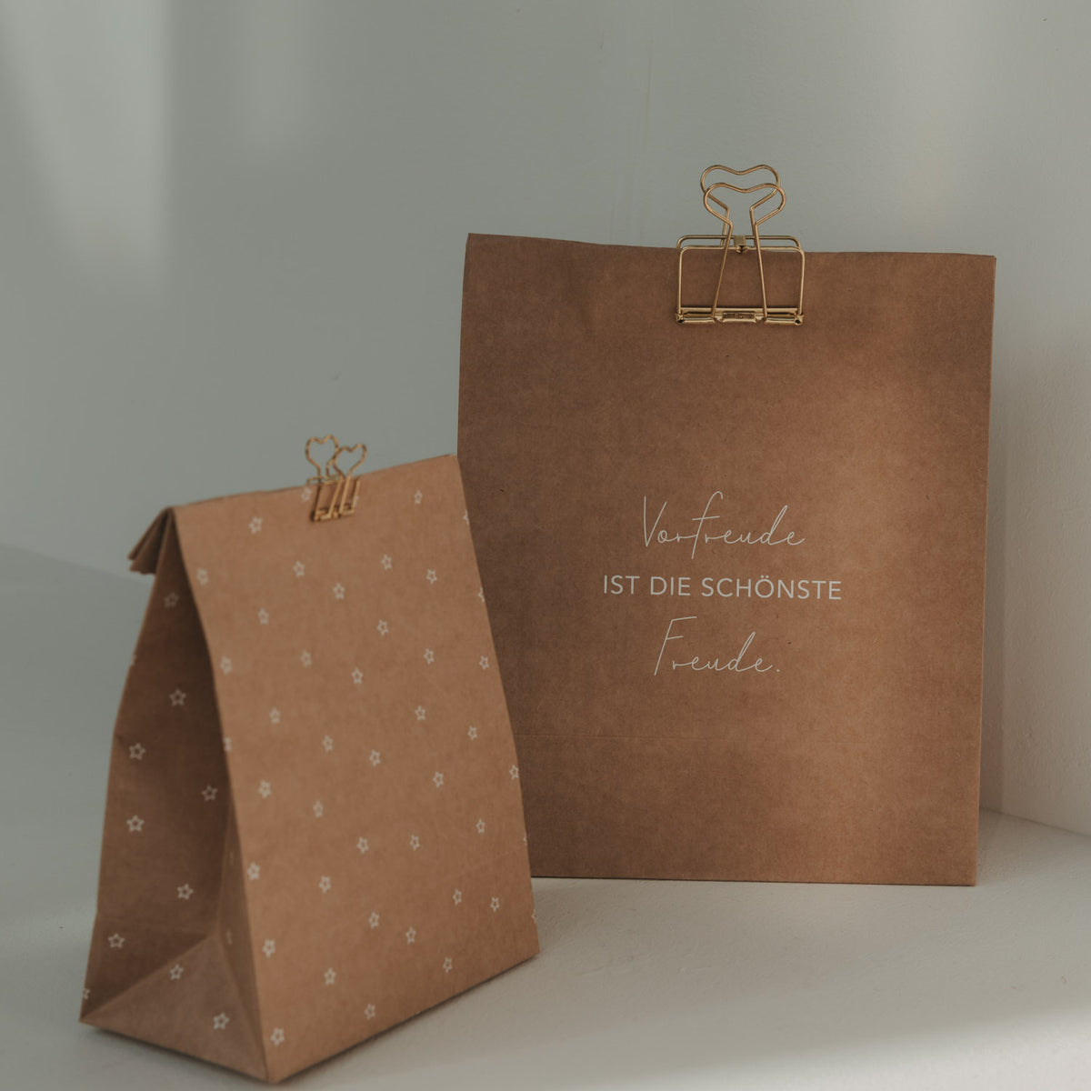 Zwei goldene Herz-Clips an braunen Geschenktüten mit Sternenmuster und dem Spruch „Vorfreude ist die schönste Freude“.