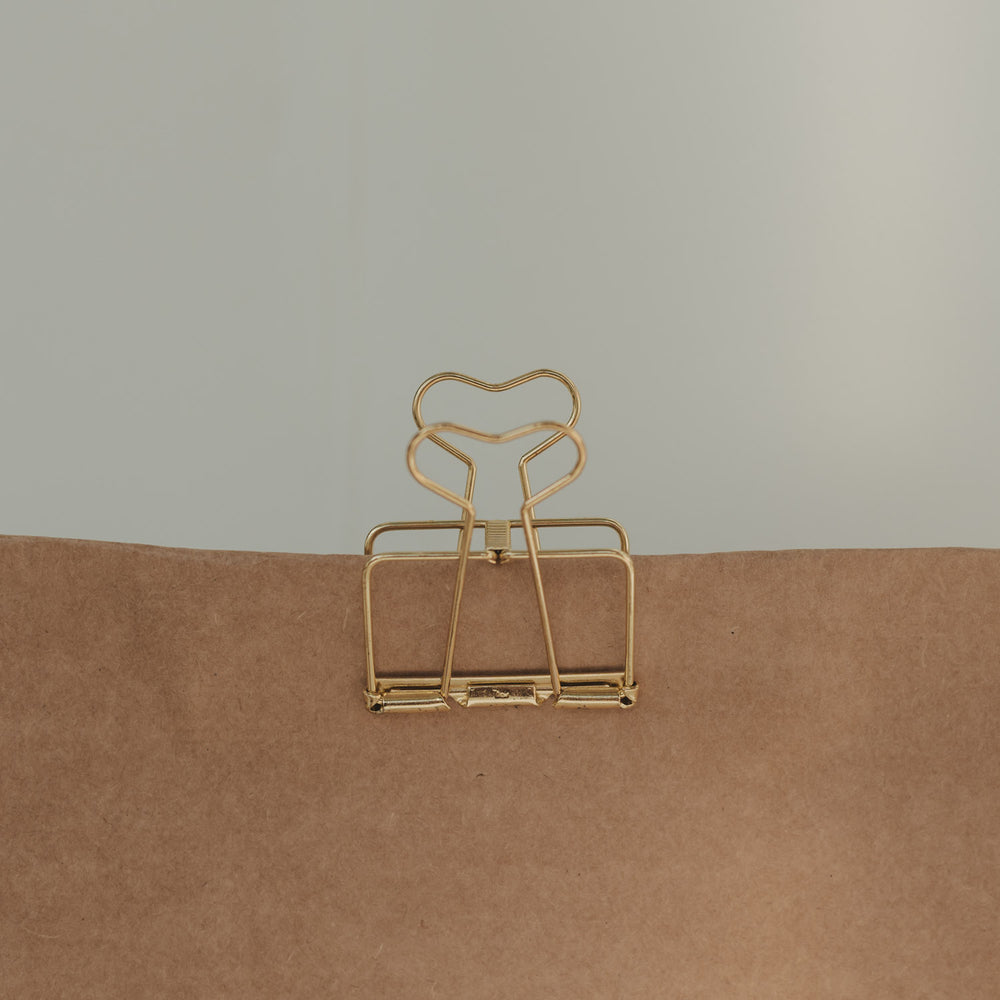 Große goldene Foldback-Klammer mit herzförmigen Griffen verschließt eine braune Geschenktüte aus Kraftpapier.
