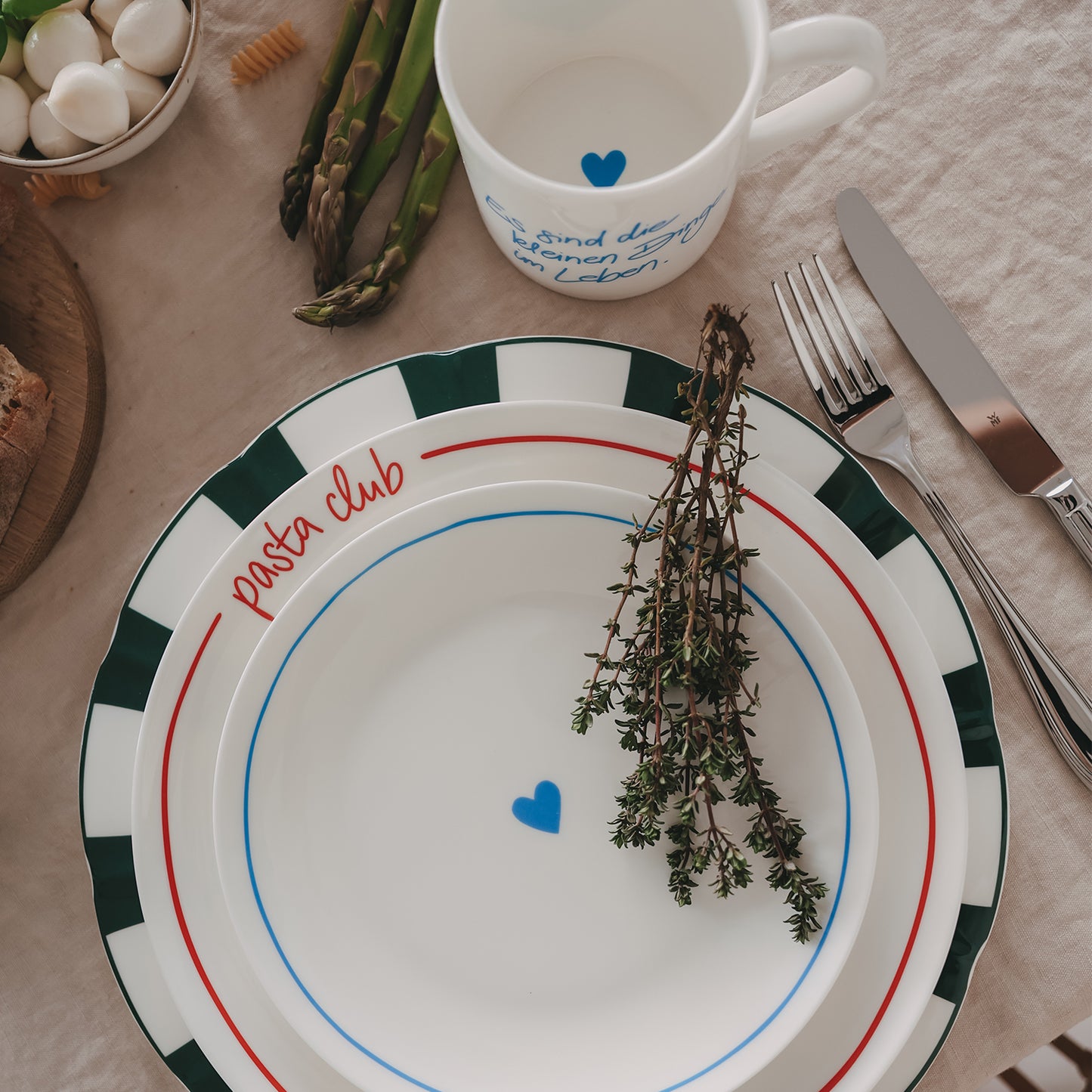 Mix & Match aus gestapelten Tellern mit Streifenmuster, Pasta-Club-Schrift und Herzmotiv, inszeniert mit frischen Zutaten wie Kräutern und Gemüse.