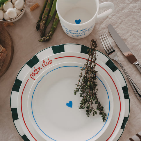 Mix & Match aus gestapelten Tellern mit Streifenmuster, Pasta-Club-Schrift und Herzmotiv, inszeniert mit frischen Zutaten wie Kräutern und Gemüse.