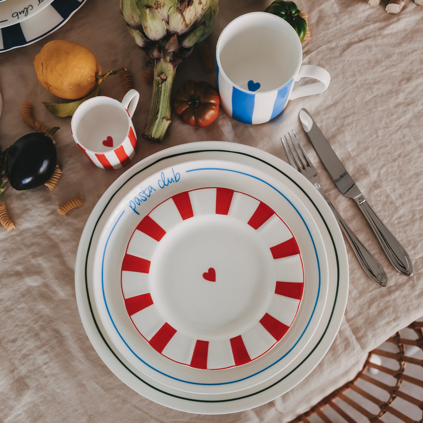 Gestapeltes Teller-Set – Frühstücksteller mit rotem Streifenrand und Herzdetail, Speiseteller Pasta Club in Hellblau, Platzset mit grünem Streifen-Rand von oben – ergänzt durch Besteck, Becher und frische Zutaten auf Leinentischdecke.