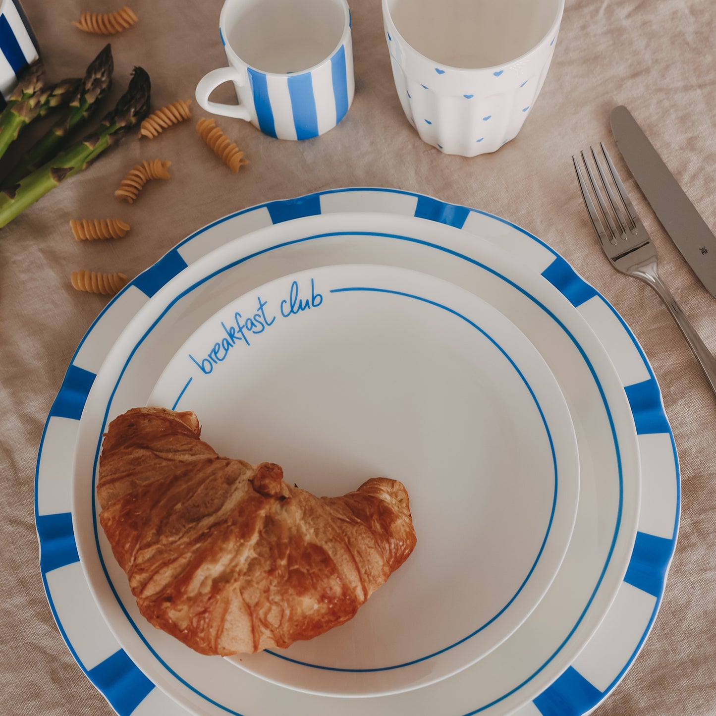 Gestapelte Teller mit blauem Streifenrand im Breakfast-Setting, kombiniert mit Croissant, Tassen, Spargel und Besteck auf natürlicher Leinentischdecke.