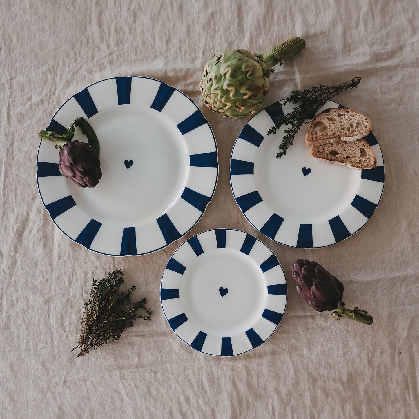 3 Teller aus Porzellan mit gewelltem Rand in verschiedenen Größen, dunkelblauen Streifen und Herzdetail, dekoriert mit Artischocken und Brot