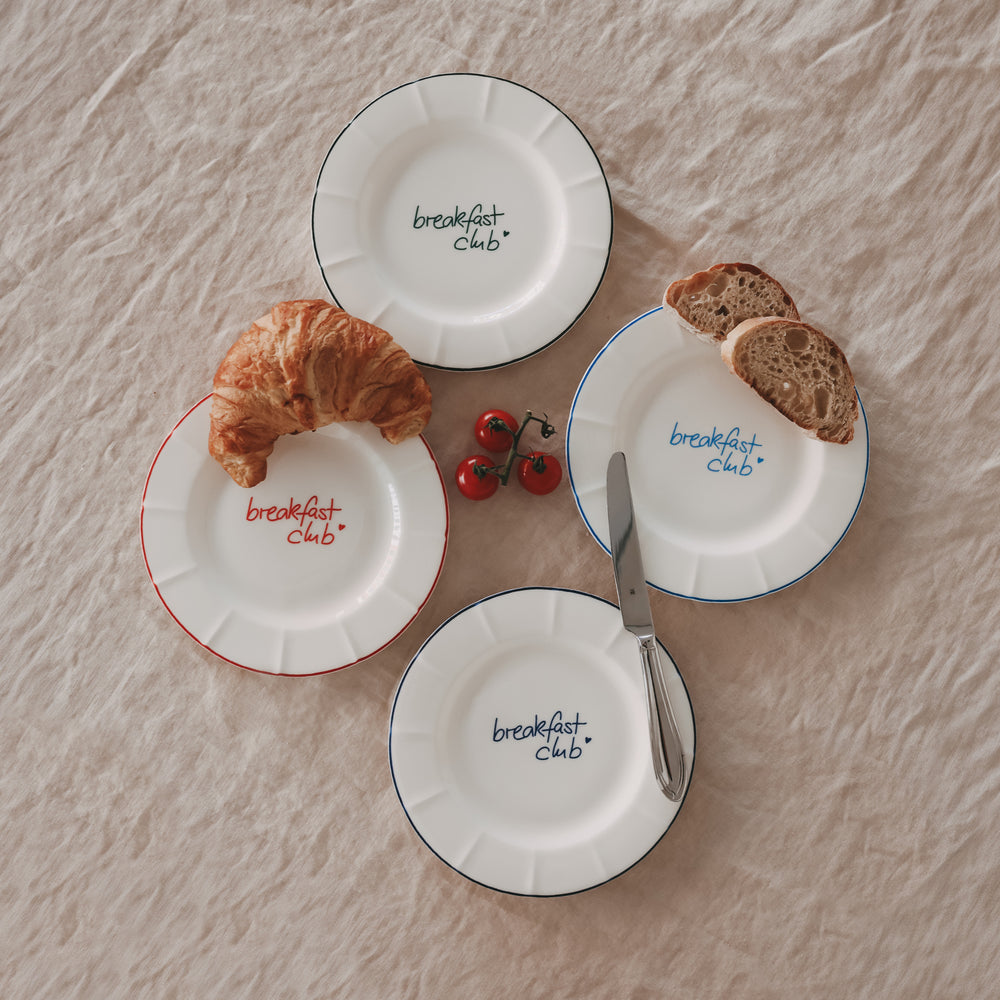 Arrangierte Frühstücksteller mit „breakfast club“ Schriftzug in verschiedenen Farben, kombiniert mit Messer, Croissant, Brot und Tomaten.