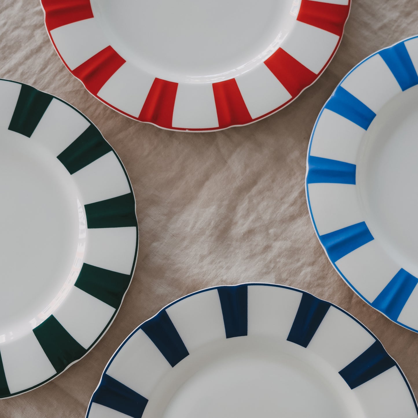 Porzellanteller mit gewelltem Rand, farbigen Streifen in Rot, Blau und Grün sowie Herzdetail, auf Leinentischdecke arrangiert.
