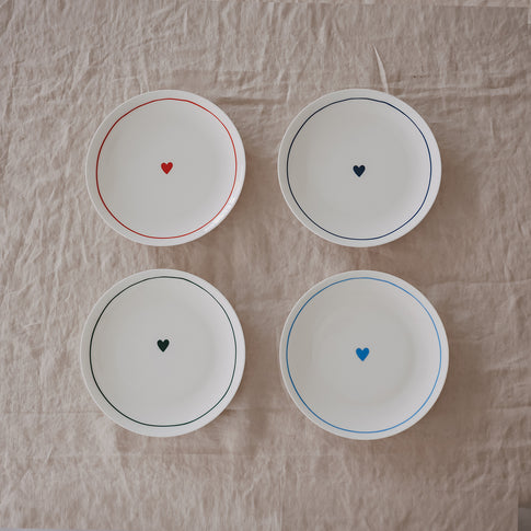 Vier runde Porzellanteller mit farbigen Rändern und kleinem Herz in der Mitte in verschiedenen Farben auf Leinentuch.