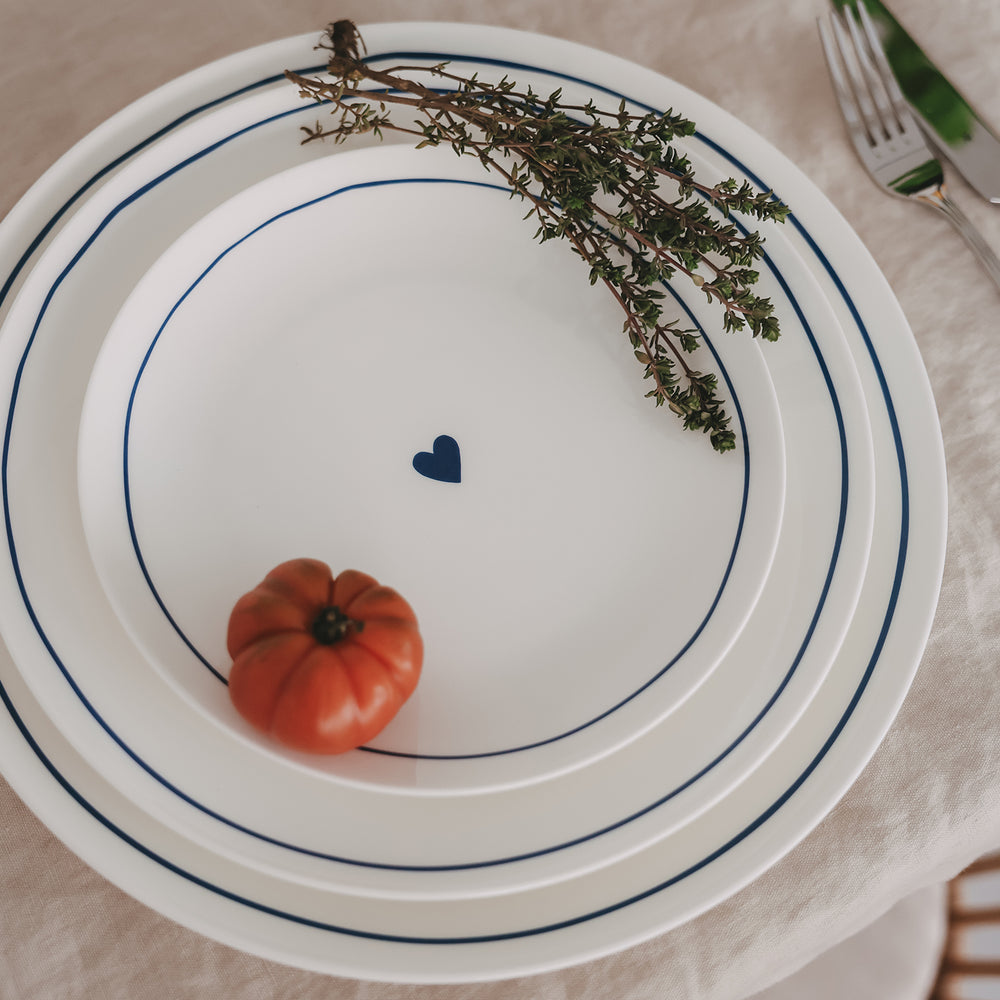 Mehrere runde Porzellanteller in 3 Größen aufeinander gestapelt – mit dunkelblauem Rand und kleinem Herz in der Mitte, dekoriert mit Thymian und Tomate.