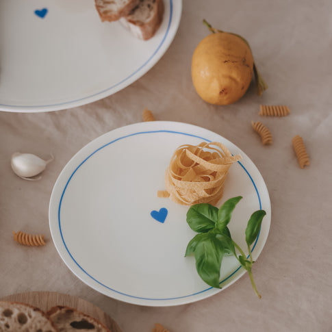 Runder Porzellanteller mit hellblauem Rand und Herzdetail, dekoriert mit Pasta und Basilikum auf Leinentuch.