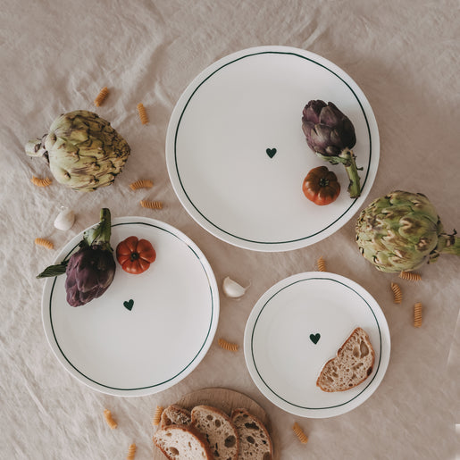 Mehrere weiße Teller mit grünem Rand und Herzmotiv, arrangiert mit Artischocken, Brot und Pasta auf Leinentuch.