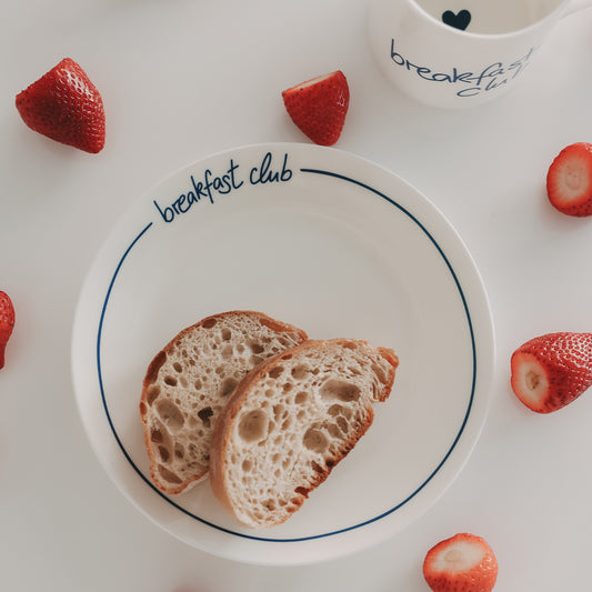 Weißer Teller mit dunkelblauem „breakfast club“-Schriftzug am Rand, darauf zwei Brotscheiben, umgeben von frischen Erdbeeren und einer Tasse im Hintergrund.