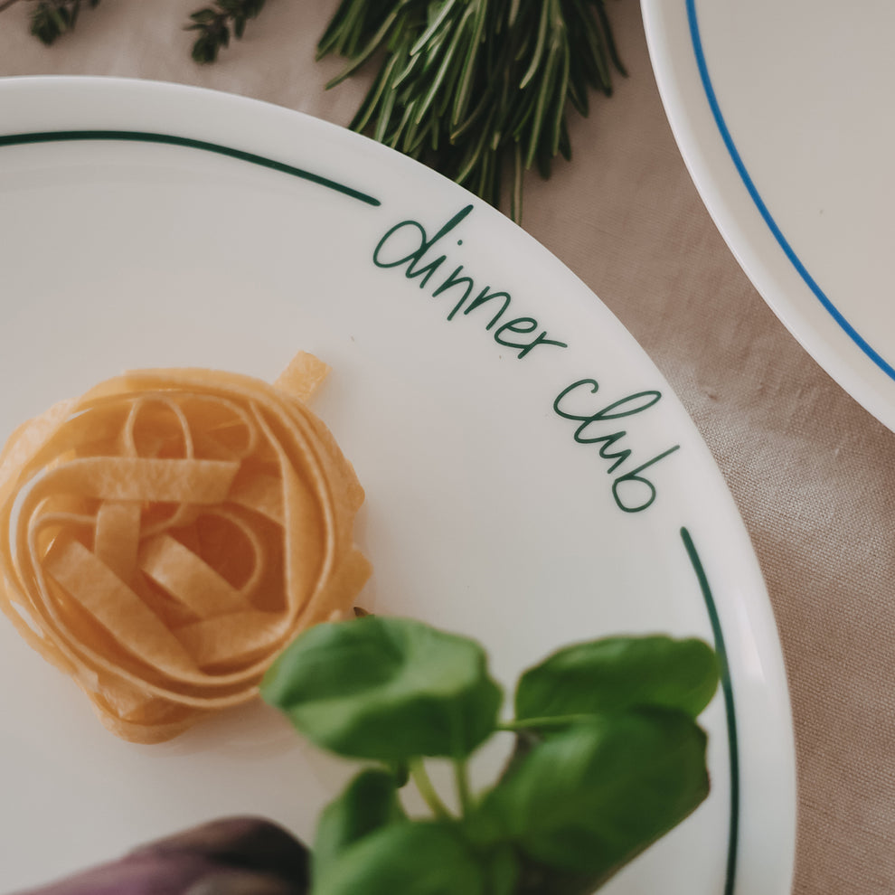 Nahaufnahme eines weißen „dinner club“-Tellers mit grünem Rand, dekoriert mit Pasta-Nest, Basilikum und Aubergine, in natürlichem Food-Styling.