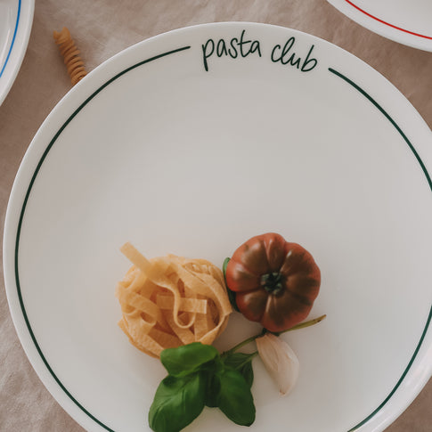 Speiseteller „pasta club“ mit grünem Rand, angerichtet mit Pasta-Nest, dunkler Tomate, Basilikum und Knoblauch auf hellem Leinentisch.