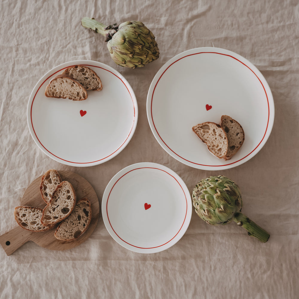 Mehrere weiße Teller mit rotem Rand und Herzmotiv, arrangiert mit Brot und Artischocken auf hellem Leinentuch.