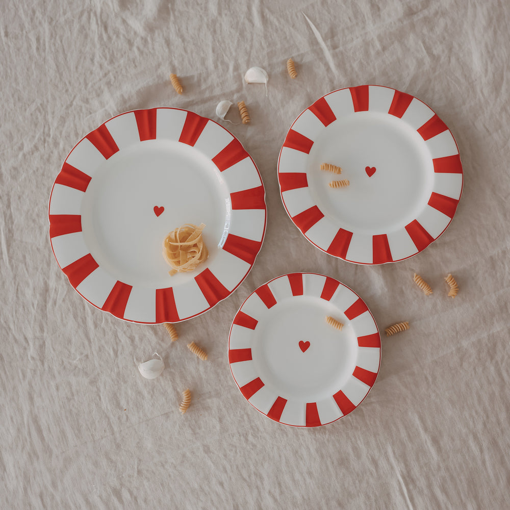 3 Arrangierte Teller mit rotem Streifenrand und Herzdetail, in verschiedenen Größen kombiniert mit Pasta und Knoblauch auf natürlicher Leinentischdecke