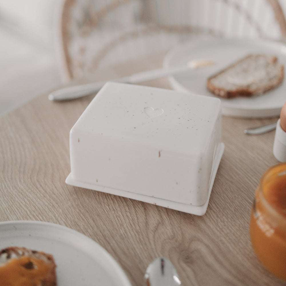 Butterdose mit Herz aus Keramik auf gedecktem Frühstückstisch mit Brot und Marmelade