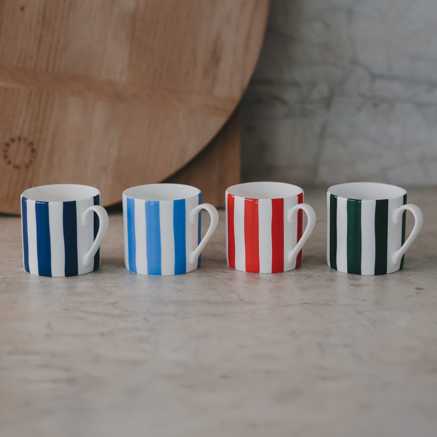 Vier kleine Tassen in einer Reihe – dunkelblau, hellblau, rot und grün – auf einer hellen Steinfläche mit Holzbrett im Hintergrund.