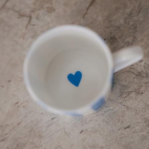 Blick in eine kleine Porzellan-Tasse mit weißem Inneren und einem kleinen blauen Herz am Boden, auf heller Steinfläche.