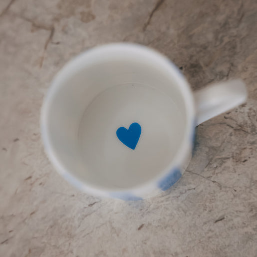 Blick in eine kleine Porzellan-Tasse mit weißem Inneren und einem kleinen blauen Herz am Boden, auf heller Steinfläche.