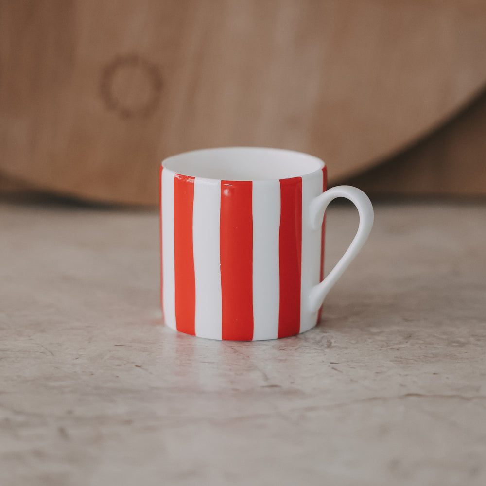 Kleine Porzellan-Tasse mit roten Streifen frontal auf heller Fläche, weicher Hintergrund mit Holzbrett.