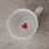 Draufsicht in die kleine weiße Tasse – am Boden ein kleines rotes Herz auf heller Steinoberfläche.
