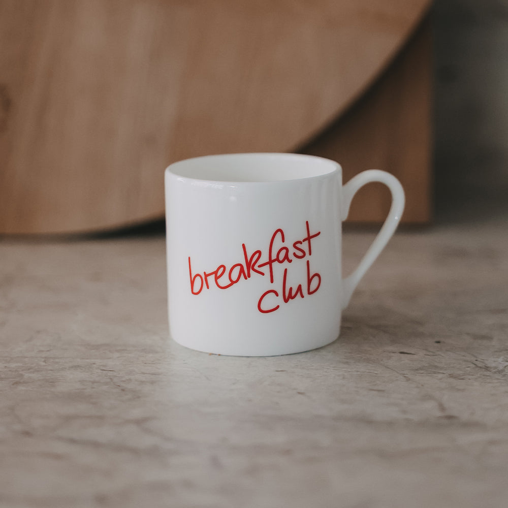 Kleine Tasse mit rotem „breakfast club“-Schriftzug, minimalistisch auf neutralem Hintergrund fotografiert.