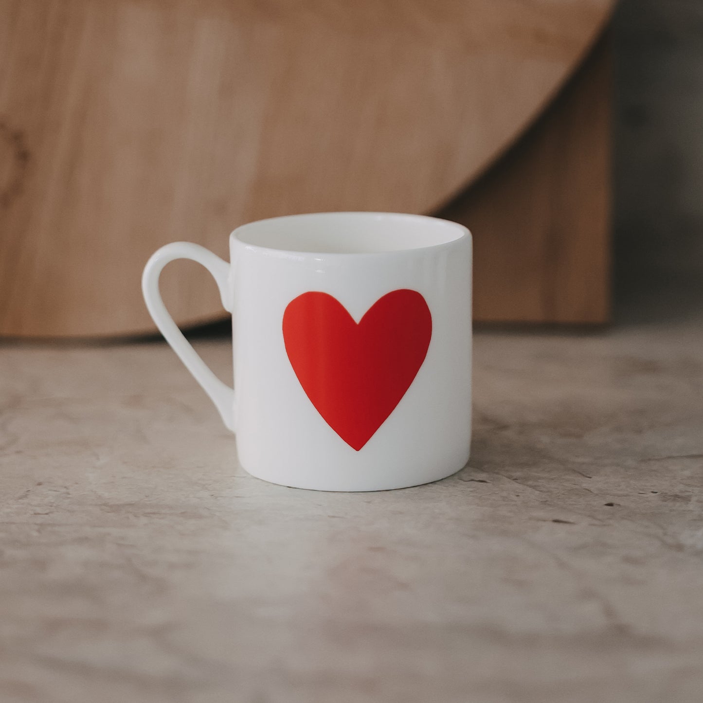Kleine weiße Tasse mit großem rotem Herz auf der Vorderseite, schlicht inszeniert vor warmem Hintergrund.