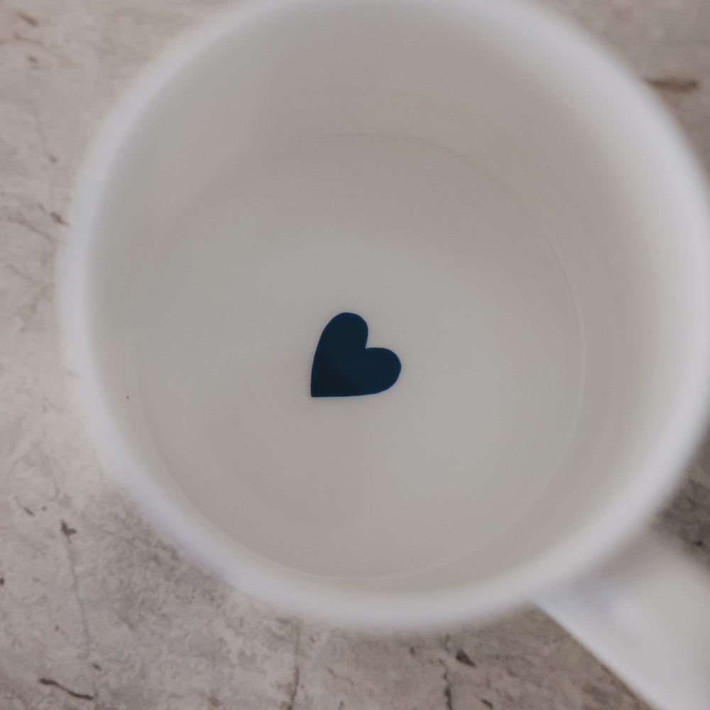 Innenansicht einer weißen Porzellantasse mit kleinem dunkelblauen Herz auf hellem Steinuntergrund