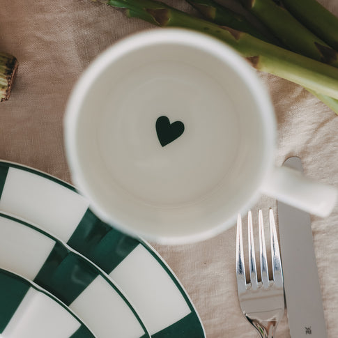 Blick in Porzellantasse mit grünem Herz am Boden, umgeben von grün gestreiftem Teller und Besteck auf hellem Leinentuch