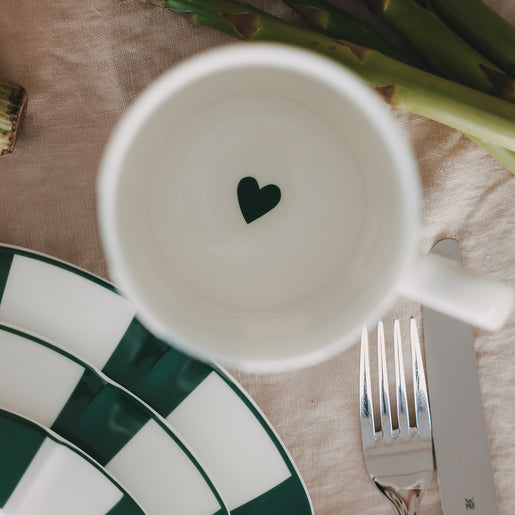 Blick in Porzellantasse mit grünem Herz am Boden, umgeben von grün gestreiftem Teller und Besteck auf hellem Leinentuch