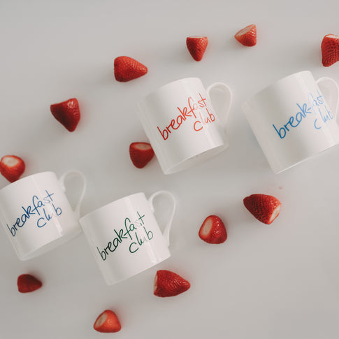 „Breakfast club“-Tassen in Rot, Blau und Grün auf weißem Hintergrund, dekoriert mit halbierten Erdbeeren