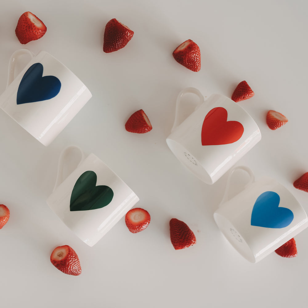 Weiße Tassen mit großen Herzmotiven in Hellblau, Blau, Grün und Rot, zwischen frischen Erdbeeren auf hellem Hintergrund