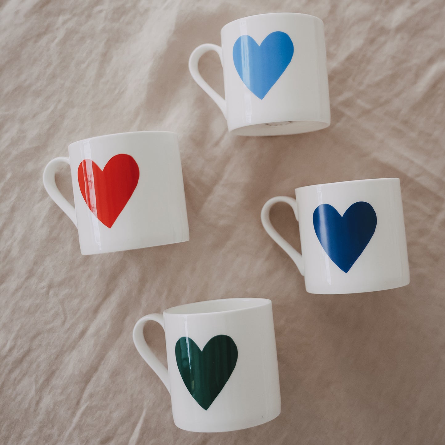 Weiße Tassen mit großen Herzmotiven in Blau, Rot und Dunkelblau locker auf Stoffuntergrund verteilt