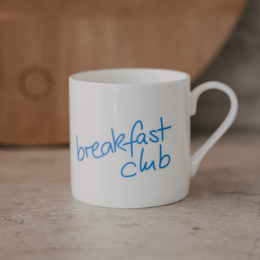 Weiße Tasse mit hellblauem „breakfast club“-Schriftzug vor hellem Untergrund