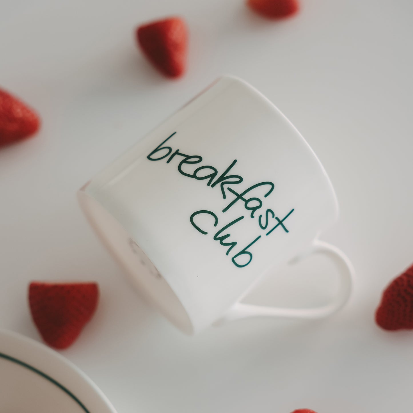 Frühstücksszene mit weißer schräg liegender Tasse mit grünem Schriftzug und Erdbeeren auf hellem Hintergrund