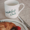 Weiße Tasse mit grünem „breakfast club“-Schriftzug neben Croissant und Messer auf gedecktem Tisch