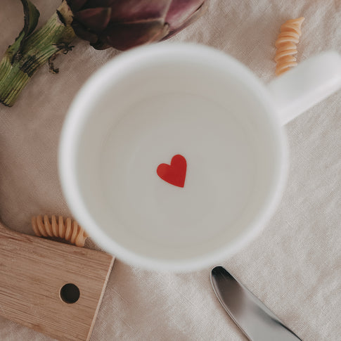 Weiße Tasse mit kleinem rotem Herz im Inneren, umgeben von Pasta und Küchenutensilien