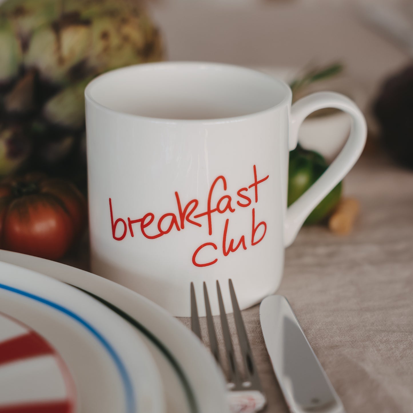Weiße Tasse mit rotem Schriftzug „breakfast club“ neben Teller und Besteck in Küchenszene