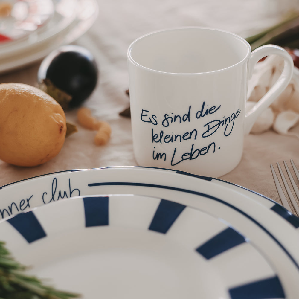 Weiße Tasse mit blauem Schriftzug „Es sind die kleinen Dinge im Leben“, dekoriert auf gedecktem Tisch mit Teller und Obst & Gemüse im Hintergrund