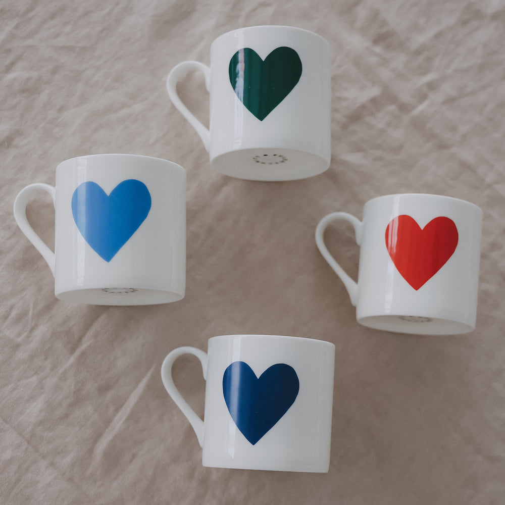 4 weiße Tassen mit großen Herzen in Blau, Grün und Rot locker auf hellem Stoff arrangiert