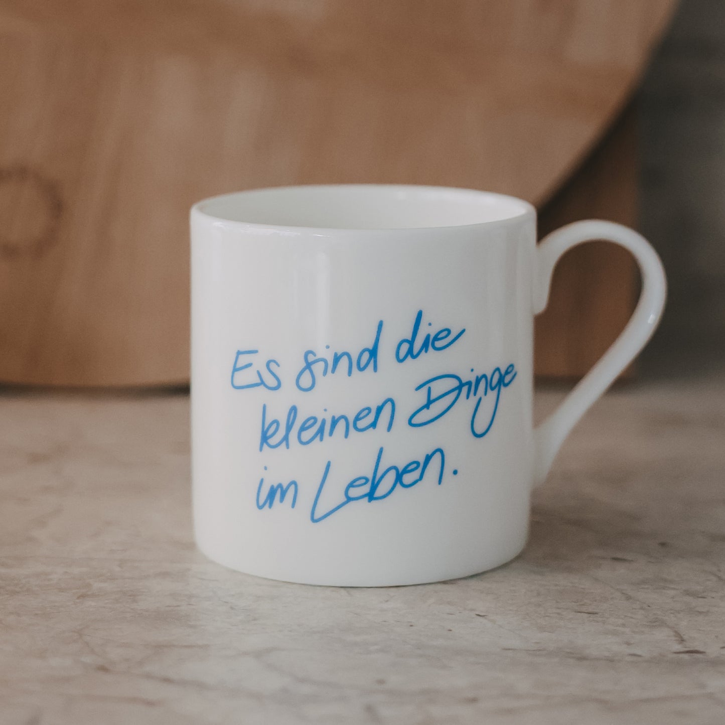 Weiße Tasse mit blauem Schriftzug „Es sind die kleinen Dinge im Leben“ vor warmem Holz-Hintergrund