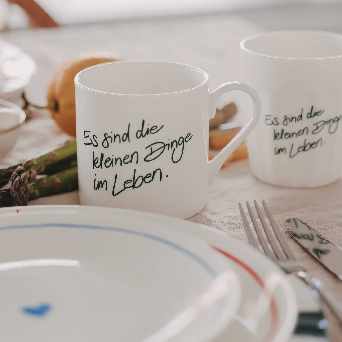 1 weiße Tasse und 1 weißer Becher mit grünem Schriftzug „Es sind die kleinen Dinge im Leben“ auf gedecktem Tisch mit Teller, Besteck und Spargel