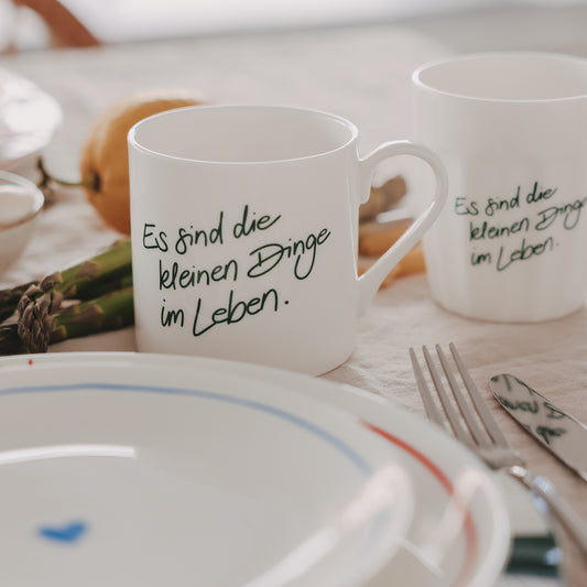 1 weiße Tasse und 1 weißer Becher mit grünem Schriftzug „Es sind die kleinen Dinge im Leben“ auf gedecktem Tisch mit Teller, Besteck und Spargel