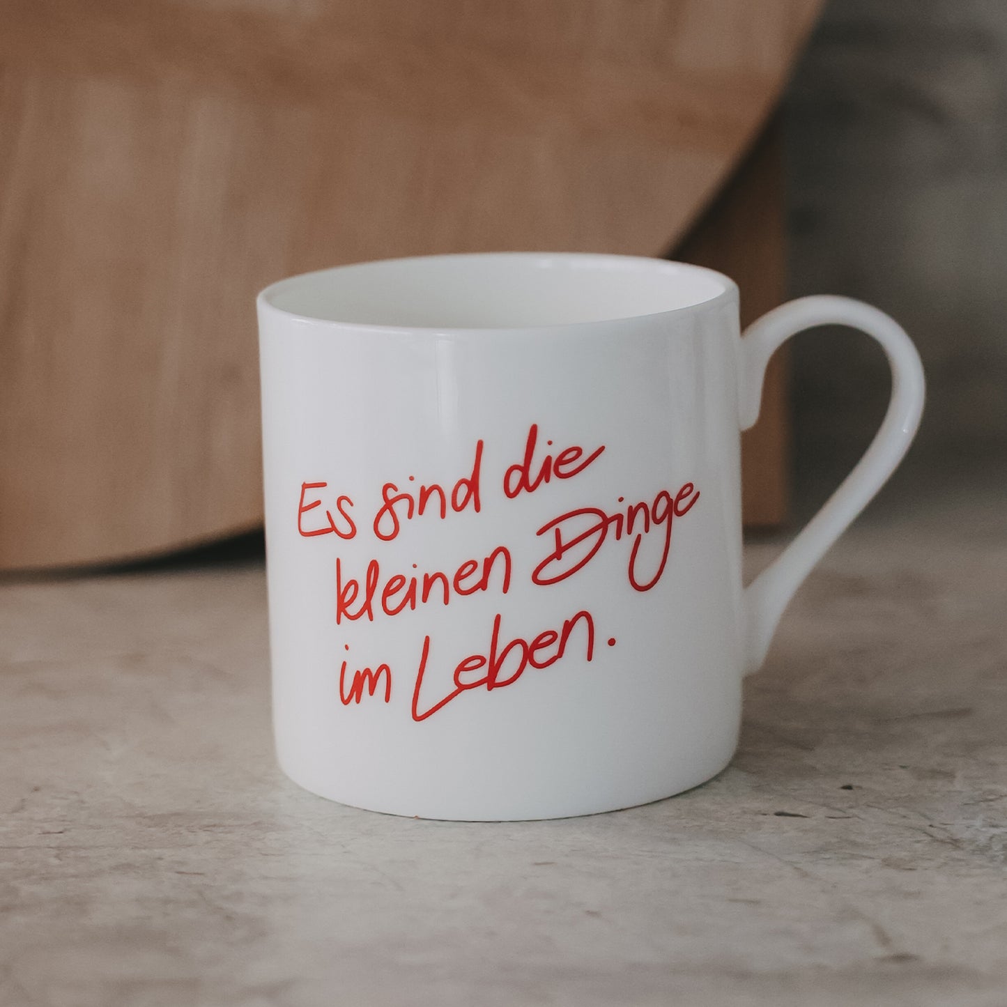Weiße Tasse mit rotem Schriftzug „Es sind die kleinen Dinge im Leben“ vor warmem Holz-Hintergrund