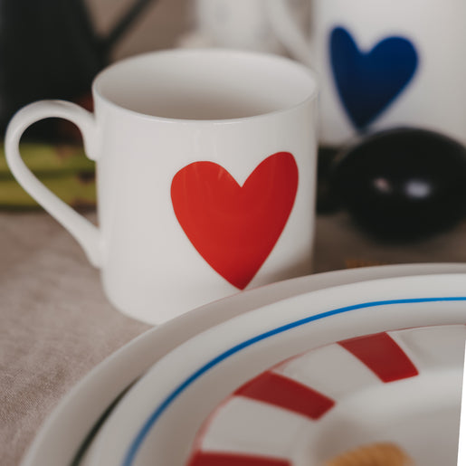 Weiße Tasse mit rotem Herzmotiv auf gedecktem Tisch, im Hintergrund weiteres Geschirr mit blauem Herz