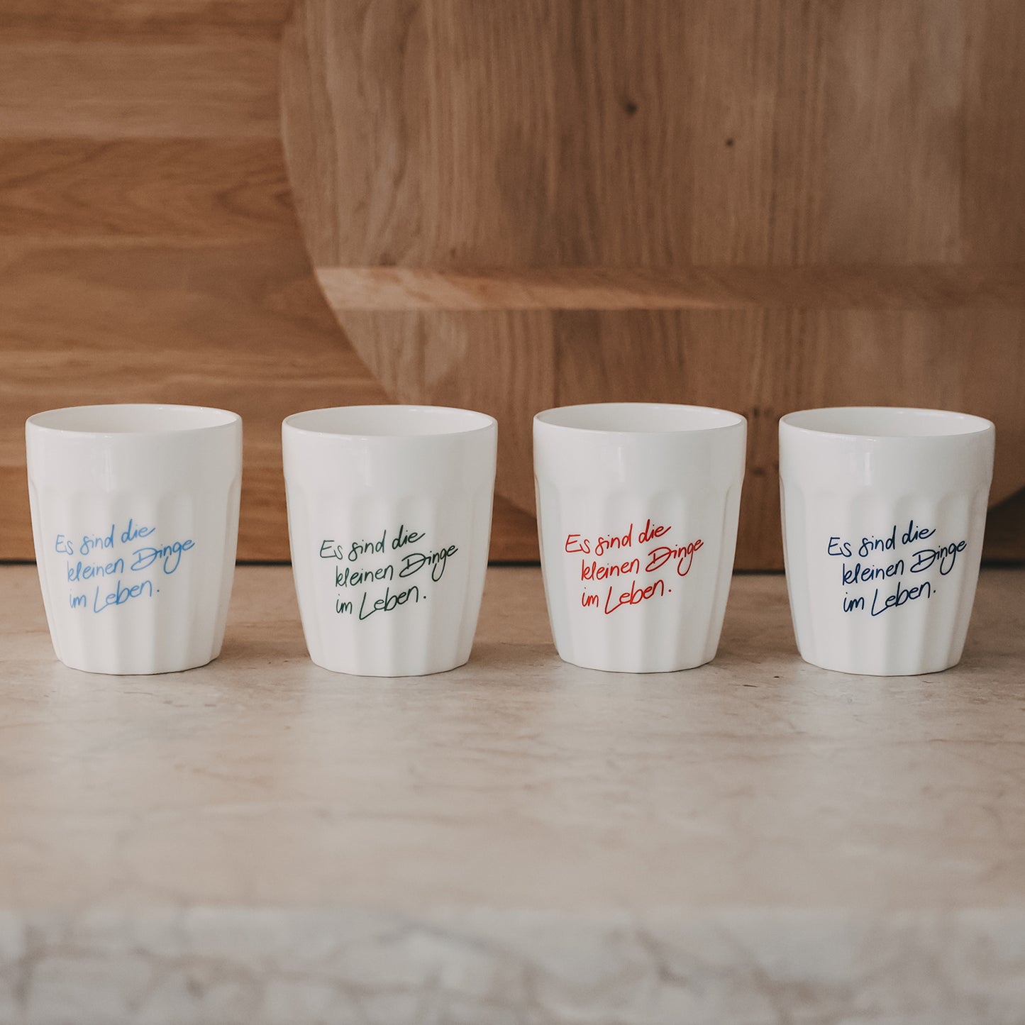 Vier weiße Keramikbecher mit Spruch „Es sind die kleinen Dinge im Leben“ in verschiedenen Farben nebeneinander